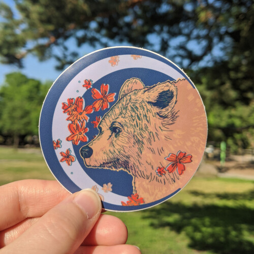 Round sticker of a bear's face and shoulders framed by flowers.